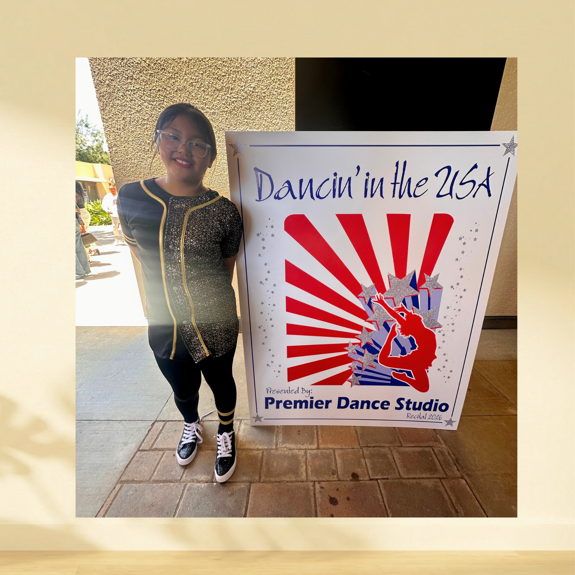 Young dancer standing beside a large red, white and blue dance recital foam board sign