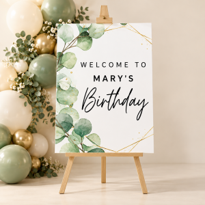 Mary's personalized birthday welcome sign on a wooden easel with eucalyptus greenery and gold accents beside sage, cream and gold balloons.