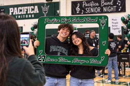 Two students posing with a green Pacifica High School Band photo frame at a senior night celebration.